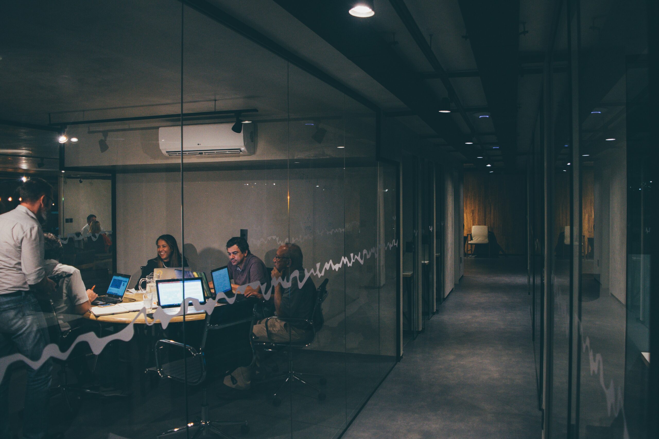 Office meeting room with several people gathered around a conference table covered with open laptops and documents, seen through a glass wall. Photo by Mario Gogh on Unsplash